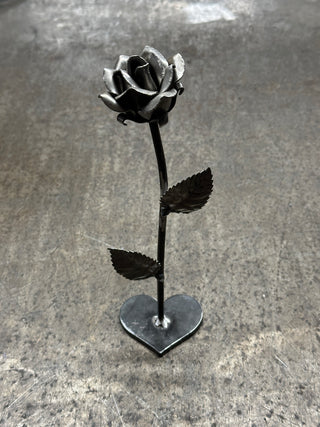 Handmade steel rose sculpture with heart shaped base displayed on workshop bench
